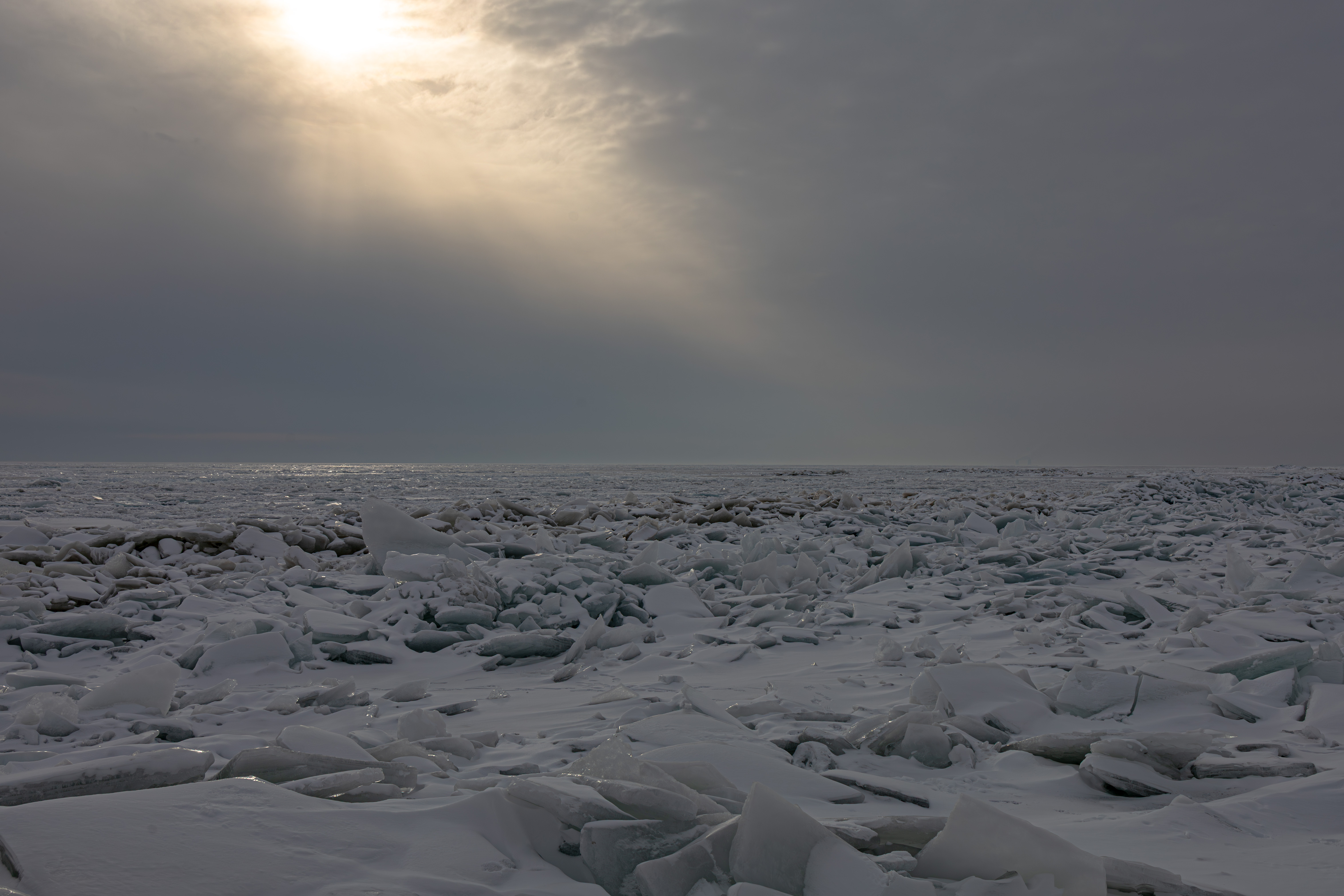 A vast expanse of snow and ice with jagged ice formations under a cloudy sky, illuminated by rays of sunlight.